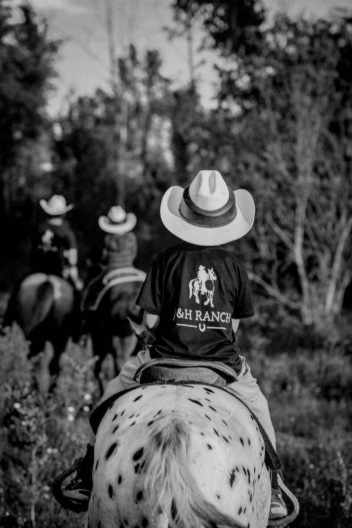 a young person at J and H Ranch is riding away from the camera wearing a cowboy hat while following two other riders